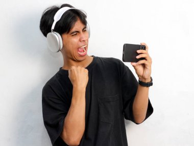 Siyah tişört giyen, elinde akıllı telefon tutan ve beyaz arka planda izole edilmiş kablosuz kulaklık kullanan Asyalı genç bir adamın portresi.