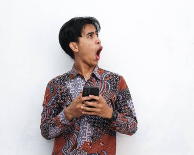 Elinde akıllı bir telefon tutarken batik takan Endonezyalı bir adamın portresi yalıtılmış beyaz bir arka planda şok olmuş bir yüzle yan tarafa bakıyor..