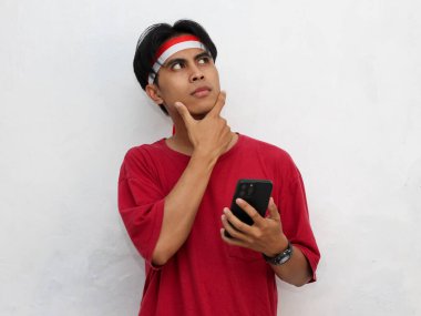 Asyalı genç bir adamın portresi düşünceli bir yüz gösteriyor, bir yandan yandan bakarken akıllı bir telefon tutuyor, çarpıcı kırmızı bir tişört ve Endonezya bandı takıyor. Endonezya Bağımsızlık Günü.