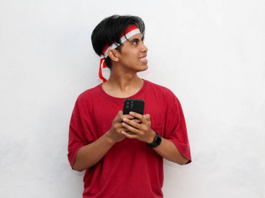 Asyalı genç bir adamın portresi mutlu bir yüz gösteriyor, yan tarafa bakarken akıllı bir telefon tutuyor, çarpıcı kırmızı bir tişört ve Endonezya bayraklı bir saç bandı takıyor. Endonezya bağımsızlık kavramı.