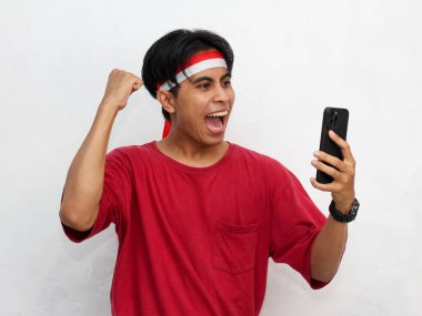 Asyalı genç bir adamın portresi heyecanlı bir yüz ifadesi, elinde akıllı bir telefon, başarılı jestler, çarpıcı kırmızı bir tişört ve Endonezya bandı takıyor..