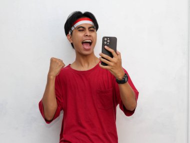 Asyalı genç bir adamın portresi heyecanlı bir yüz ifadesi, elinde akıllı bir telefon, başarılı jestler, çarpıcı kırmızı bir tişört ve Endonezya bandı takıyor..