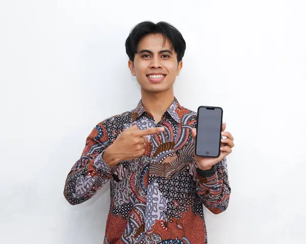 stock image Portrait of a funny wow surprised Indonesian man wearing batik while holding a smartphone and pointing at the smartphone with a blank screen, presenting a sales discount promo offer.