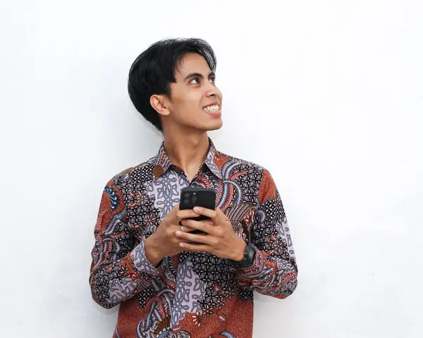 stock image Portrait of a smiling handsome Indonesian man wearing batik, holding a smartphone and looking away on an isolated white background.