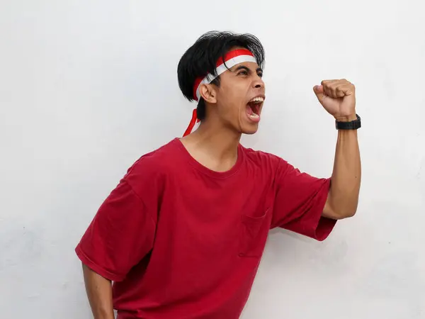 stock image Portrait of an Asian young man, wearing a striking red t-shirt and headband with the Indonesian flag. Raising fist enthusiastically, celebrating Indonesia's independence August 17.