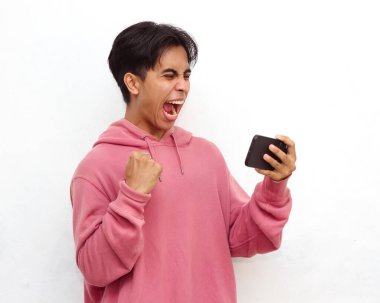 Çok heyecanlı genç Asyalı bir adamın portresi, pembe kapüşonlu, akıllı telefon tutan ve oyun oynayan, kazanan hareketi beyaz arka planda izole eden.