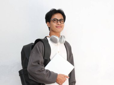 Gözlükleri, kulaklıkları ve sırt çantasıyla izole edilmiş beyaz arka planı olan mutlu Asyalı genç bir adam.