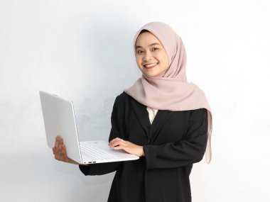 Genç Müslüman kadın tesettür takıyor ve dizüstü bilgisayarını beyaz arka planda tutuyor.