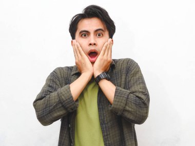 Şok olmuş yeşil fanila gömlekli Asyalı adam beyaz arka plandaki kameraya bakıyor.    