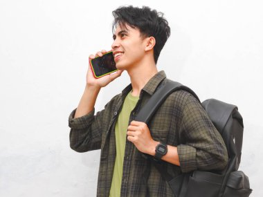 Yeşil flanel gömlekli Asyalı adam beyaz arka planda sırt çantası ve telefonla poz veriyor.    