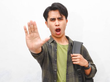 Yeşil flanel gömlekli Asyalı adam sırt çantalı ve beyaz arka planda dur işareti gösteriyor.     