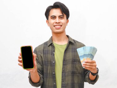 Yeşil flanel gömlekli Asyalı adam beyaz arka planda para ve telefon tutuyor.  