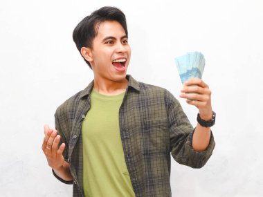 Yeşil flanel gömlekli Asyalı adam beyaz arka planda para tutuyor.  