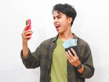 Üzerinde para ve telefon olan yeşil fanila gömlekli Asyalı adam beyaz arka planda cüzdan tutuyor.  