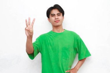 Gülümseyen ve kendinden emin Endonezyalı, yeşil tişört giyen, elini kaldıran ve kameraya parmakları olan 3 numarayı gösteren.