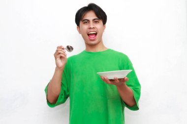 Yeşil tişörtlü neşeli yetişkin bir Asyalı elinde boş bir tabakla poz verirken beyaz arka planda izole edilmiş aç bir yüz gösteriyor.