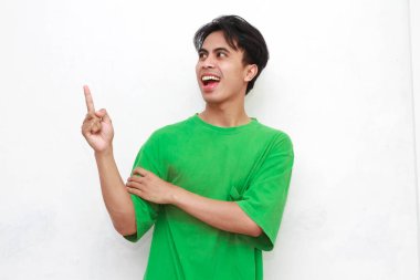 Yeşil tişörtlü neşeli yetişkin Asyalı adam beyaz arka planda izole edilmiş boş bir bölgeyi işaret ediyor.
