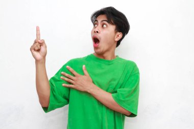 Yeşil tişörtlü neşeli yetişkin Asyalı adam beyaz arka planda izole edilmiş komik bir ifadeyle boş bölgeyi işaret ediyor.