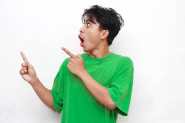 Yeşil tişörtlü neşeli yetişkin Asyalı adam beyaz arka planda izole edilmiş komik bir ifadeyle boş bölgeyi işaret ediyor.