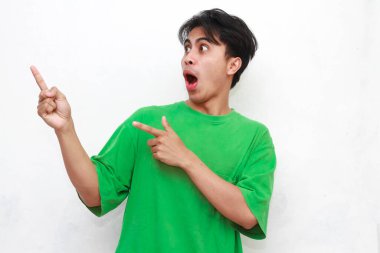 Yeşil tişörtlü neşeli yetişkin Asyalı adam beyaz arka planda izole edilmiş komik bir ifadeyle boş bölgeyi işaret ediyor.