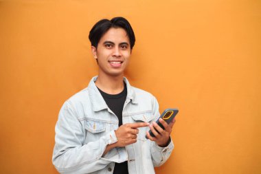 Akıllı telefonu işaret eden mutlu Asyalı genç adam. Akıllı telefonu tutan neşeli Asyalı genç adam. Sarı arka planda izole edilmiş bir tarafa bakıyor..