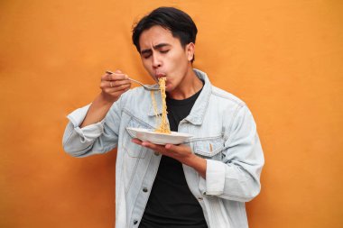 Sarı gömlekli genç bir adam, parlak sarı bir arka planda çatalla bir tabak erişte yiyor..