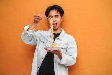 Sarı gömlekli genç bir adam, parlak sarı bir arka planda çatalla bir tabak erişte yiyor..