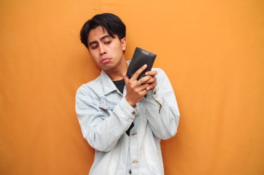 Zavallı Asyalı adam boş cüzdana bakıyor sarı arka planda izole edilmiş tuhaf bir yüz gösteriyor.