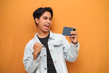 Mobil video oyunu uygulamasında akıllı telefon oyunu oynayan heyecanlı Asyalı genç oyuncu ya da sarı arka planda izole edilmiş dijital video akışını izleyen.