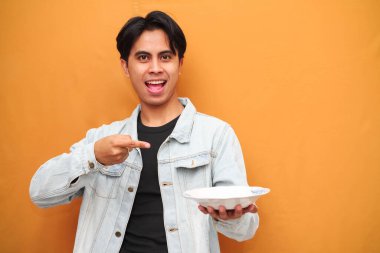 Heyecanlı Asyalı genç adam, aç suratlı, çalışma alanı olan boş bir tabak taşıyor ve sarı arka planda elini işaret ediyor.