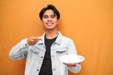 Heyecanlı Asyalı genç adam, aç suratlı, çalışma alanı olan boş bir tabak taşıyor ve sarı arka planda elini işaret ediyor.