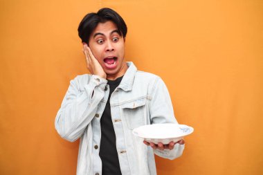 Heyecanlı Asyalı genç adam, aç suratlı, çalışma alanı olan boş bir tabak taşıyor ve sarı arka planda elini işaret ediyor.