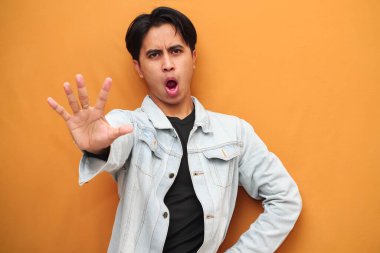 Asian young man with serious face posing making stop sign with hand, Angry Asian young man doing stop gesture isolated on yellow background, defensive gesture, suitable for advertising purposes. clipart