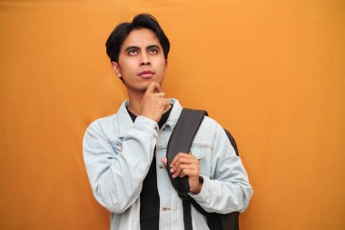 Sırt çantası taşıyan Asyalı bir öğrencinin portresi. Eli çenesinde. İzole edilmiş sarı arka planda.