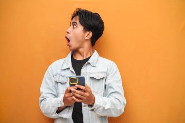 Asyalı genç adam elinde akıllı telefonuyla yan tarafı işaret ederken hayrete düştü. Asyalı komik suratlı, sarı arka planda izole edilmiş akıllı telefona bakan adam..