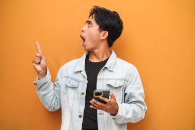 Asyalı genç adam elinde akıllı telefonuyla yan tarafı işaret ederken hayrete düştü. Asyalı komik suratlı, sarı arka planda izole edilmiş akıllı telefona bakan adam..