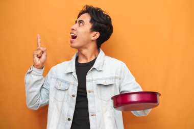 Asyalı genç bir adam kızartma tavası tutuyor, Asyalı bir adam elinde kırmızı tavayla elini gösterirken sarı arka planda izole edilmiş, yemek reklamı için mükemmel bir yüz ifadesi sergiliyor.