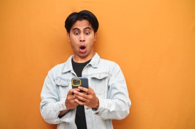 Asyalı genç adam elinde akıllı telefonuyla yan tarafı işaret ederken hayrete düştü. Asyalı komik suratlı, sarı arka planda izole edilmiş akıllı telefona bakan adam..