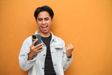 Heyecanlı mutlu Asyalı genç adam piyango kazanan akıllı telefon oyunu oynayarak mutlu oluyor, bahis oynuyor, cep telefonuyla mobil uygulama mesajı alıyor sarı arka planda izole edilmiş..
