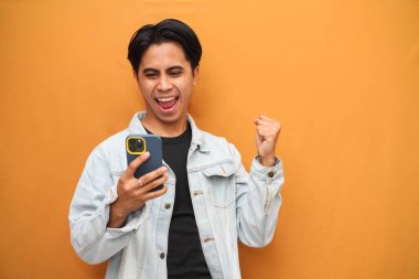 Heyecanlı mutlu Asyalı genç adam piyango kazanan akıllı telefon oyunu oynayarak mutlu oluyor, bahis oynuyor, cep telefonuyla mobil uygulama mesajı alıyor sarı arka planda izole edilmiş..
