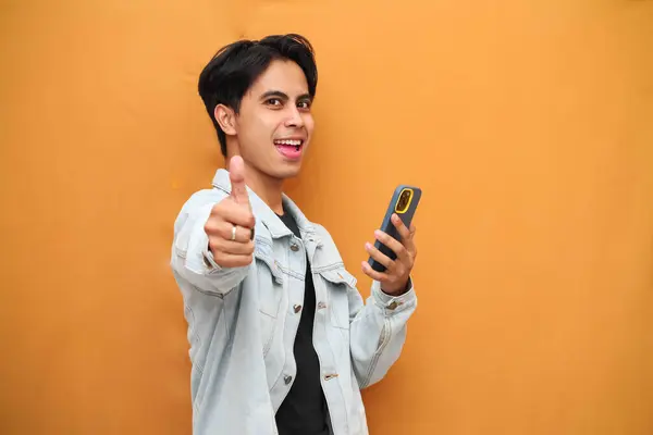 stock image happy young asian man holding smartphone while pointing at smartphone, cheerful young asian man holding smartphone while looking to the side isolated on yellow background.