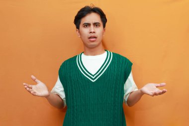 Yeşil kazak giyen genç Asyalı adam, elini kaldırıp turuncu arka planda kafası karışmış bir şekilde soru sorarak kafasını karıştırıyor.