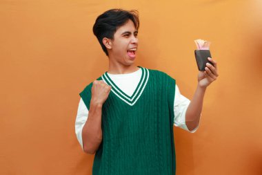 Mutlu, heyecanlı, yeşil kazaklı genç Asyalı adam turuncu arka planda parası olan bir cüzdan taşıyor.