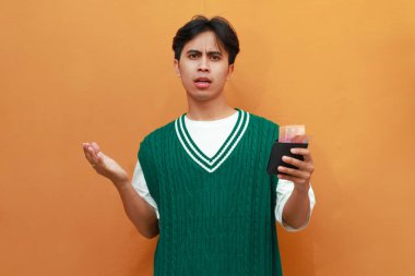 Mutlu, heyecanlı, yeşil kazaklı genç Asyalı adam turuncu arka planda parası olan bir cüzdan taşıyor.