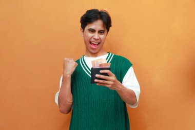 Mutlu, heyecanlı, yeşil kazaklı genç Asyalı adam turuncu arka planda parası olan bir cüzdan taşıyor.