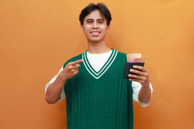 Mutlu, heyecanlı, yeşil kazaklı genç Asyalı adam turuncu arka planda parası olan bir cüzdan taşıyor.