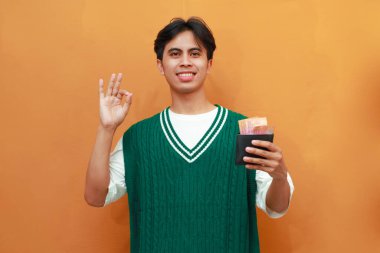 Mutlu, heyecanlı, yeşil kazaklı genç Asyalı adam turuncu arka planda parası olan bir cüzdan taşıyor.