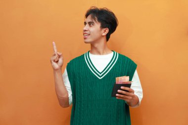 Mutlu, heyecanlı, yeşil kazaklı genç Asyalı adam turuncu arka planda parası olan bir cüzdan taşıyor.