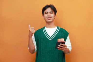 Mutlu, heyecanlı, yeşil kazaklı genç Asyalı adam turuncu arka planda parası olan bir cüzdan taşıyor.