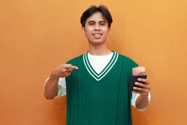 Mutlu, heyecanlı, yeşil kazaklı genç Asyalı adam turuncu arka planda parası olan bir cüzdan taşıyor.